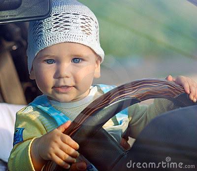 A baby sitting in a car, hands on the steering wheel